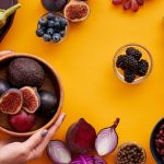 Colorful arrangement of fruits and vegetables on a yellow background, featuring figs and eggplants.